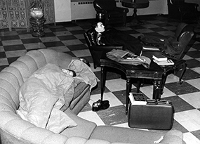 Two male students lay asleep on couches in a lounge of a college building underneath a coat and a blanket with a suitcase, shoes and a book nearby.